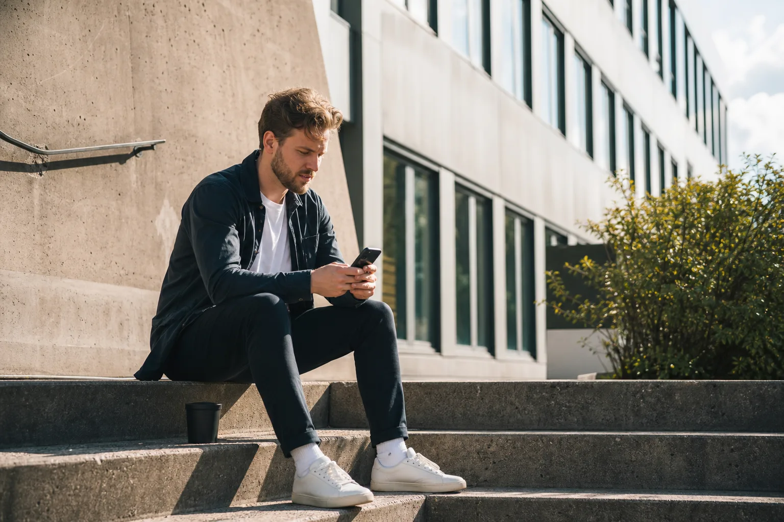 Man i trettioårsåldern sitter på en trappa utanför ett kontor på lunchrasten med en kaffemugg bredvid sig och mobilen i handen