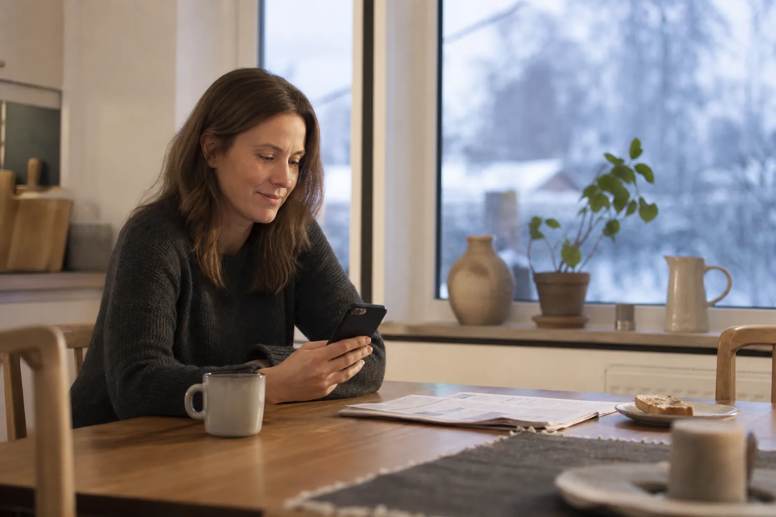 Kvinna i fyrtioårsåldern sitter vid ett köksbord i morgonljus med telefonen i handen, en halvdrucken kaffekopp och en tidning bredvid
