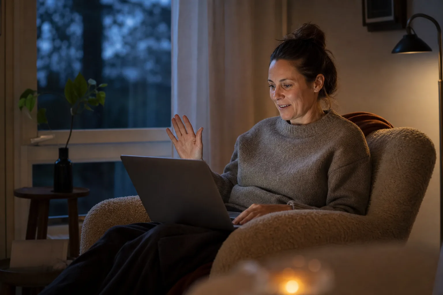 En person sitter i en fåtölj vid fönstret på kvällen med en bärbar dator i knäet, redo att föreslå ett videosamtal